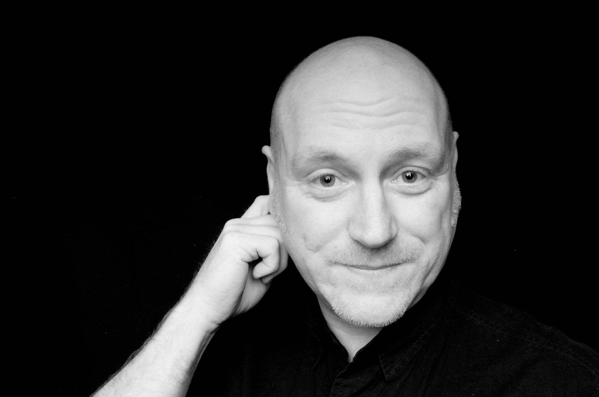 Black and white studio portrait of Elliott Richmond against a black background, hand raised to his ear